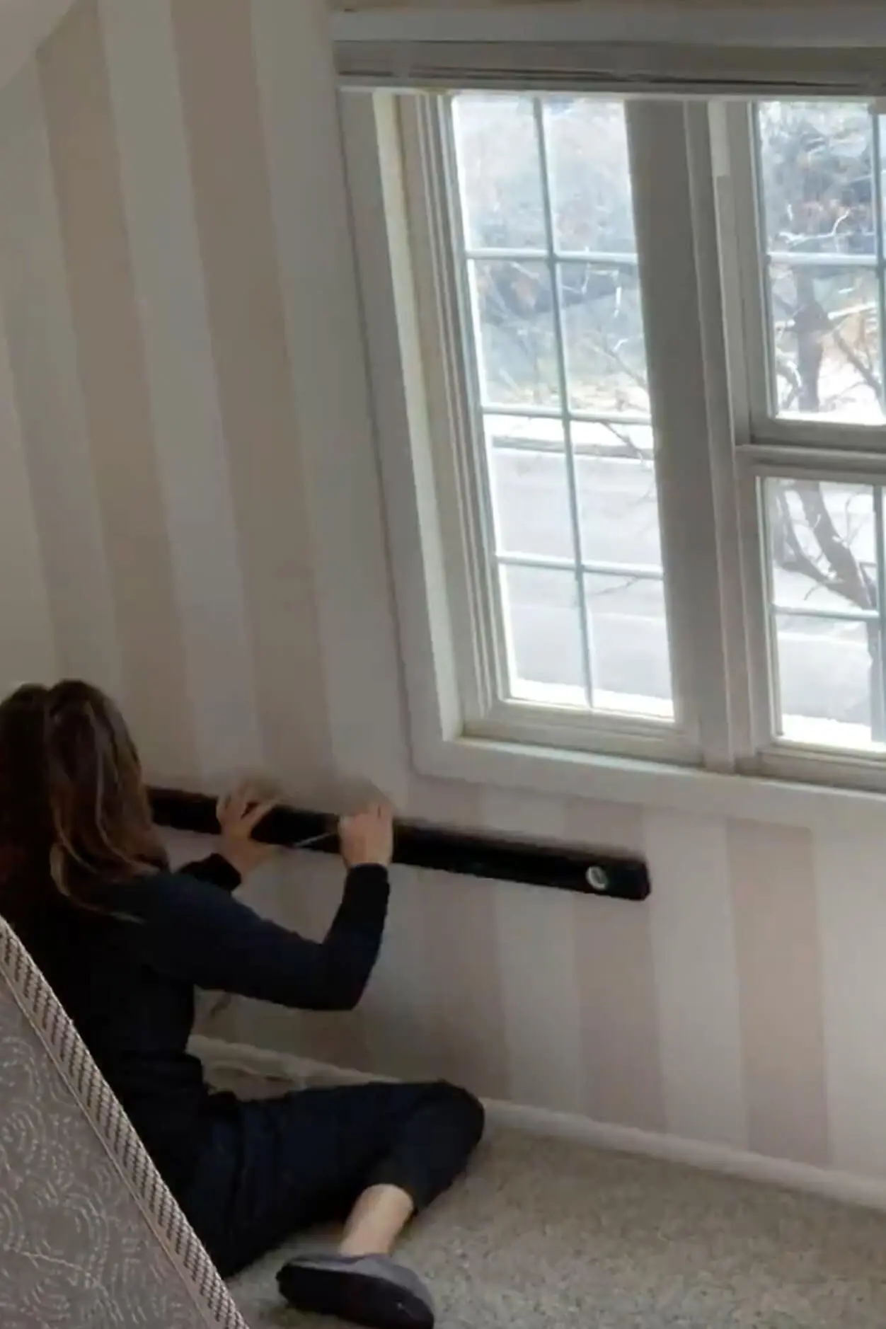 Woman using a level to draw horizontal guidelines on a striped bedroom wall in preparation for painting a pattern.